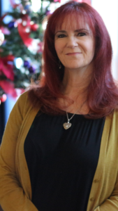 A 55 year old woman, red hair, gold sweater, black shirt, silver heart pendant necklace, dark eyes, happy smile, blurred colors of pink and green.